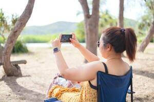 Woman wearing dark glasses smiling and taking a photo with smartphone in the park.