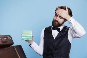 Doorman displays box of pain relievers for headache while posing against blue background. Male bellhop holding medication pack and seeking to ease migraine with medical products in studio. photo