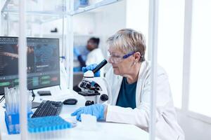 Senior scientist doing medical analysis and engineering using microscope. Elderly researcher carrying out scientific research in a sterile lab using a modern technology. photo