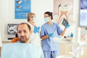 estomatólogo médico y ortodoncista asistente examinando dientes radiografía comprobación somatología problema mientras enfermo paciente esperando en dental silla. hombre preparando para cirugía cita foto