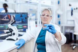 Senior scientist in protective gear holding sample during clinic study giving expertise. Elderly researcher in sterile lab looking on microscope slide wearing lab coat. photo