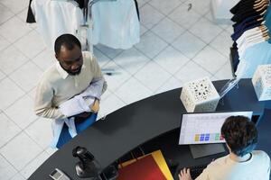 Clothing store worker using cash register software on computer while buyer standing in line at checkout top view. Shopping center employee cashier working at counter desk while customer waiting photo