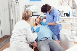 Sick man sitting on dental chair with open mouth while medical nurse with face mask and gloves analyzing teeth health during stomatology consultation. Hospital team working in stomatological clinic photo