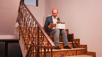 Fired businessman sitting on stairs in corporate finance company holding box of personal employee stuff. Group of professional successful businesspeople working in modern financial building. photo