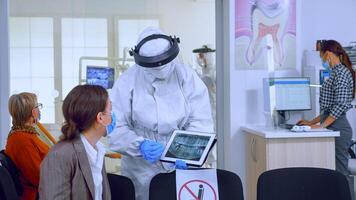 Stomatologist in protective suit pointing on digital x-ray of tooth explaining to patient treatment using tablet in covid-19 pandemic. Medical team wearing face shield, coverall, mask and gloves. photo