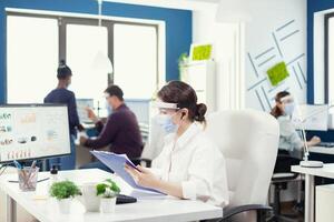 Business person using wireless headphones to talk with colleagues from another department. Employees with visors working in corporate company workspace respecting social distance analysing data and graphs. photo