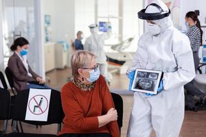 Dentist doctor with face shield pointing on tablet display explaining dental x-ray to senior patient during global pandemic. Nurse wearing protection suit, overall, mask and gloves, new normal. photo