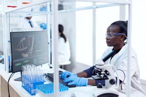 African chemist researcher typing information on computer from experimental drug trial. Black healthcare scientist in biochemistry laboratory wearing sterile equipment. photo