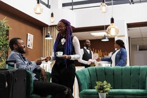 Overjoyed satisfied African American man tourist giving cash money to waitress girl while sitting in hotel lobby with luggage. Traveler buying coffee while waiting for guest check-in procedure photo