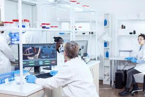 Elderly virologist working on computer with virus on display in modern facility. Senior scientist in pharmaceuticals laboratory doing genetic research wearing lab coat with team in the background. photo