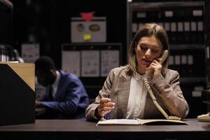 Woman detective working at criminal investigation, discussing crime scene evidence with inspector using landline phone. Police officer analyzing victim report, wanting to bring suspect to justice. photo