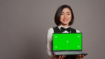 Asian hotel concierge holding pc with greenscreen on camera, presenting isolated mockup template on screen. Receptionist showing laptop with blank chromakey layout on display. Camera B. photo