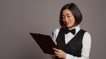 Asian hotel concierge creating a checklist of registrations, working on online reservations and bookings in studio. Receptionist looking at clipboard papers over grey background. Camera B. photo