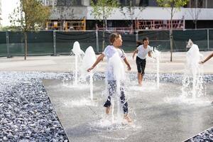 alegre joven adolescente niña en ciudad fuente, niña en mojado ropa es teniendo divertido y disfrutando el frio verano agua, antecedentes ciudad arquitectura. foto