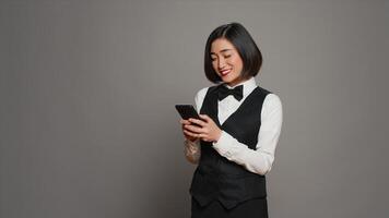 Hospitality industry worker using smartphone app over grey background, looking at online information to confirm room reservations. Asian receptionist browses websites. Camera A. photo