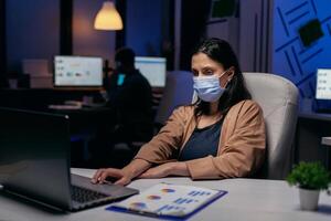 Stressed caucasian young entrepreneur doing overtime wearing face mask . Woman following social distancing rules due to coroanvirus pandemic while working late hours at the office. photo