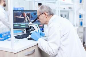 Senior caucasian scientist microbiology laboratory conducting virus analysis looking through microscope. Chemist researcher in sterile lab doing experiments for medical industry using modern technology. photo