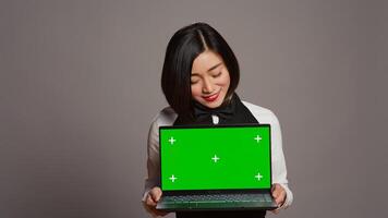 Asian server presenting greenscreen display on laptop in studio, wearing uniform with apron and bow. Restaurant hostess holding pc with blank mockup template, copyspace layout. Camera B. photo