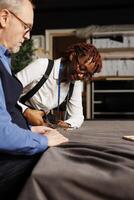 African american seamstress using scissors to cut refined fabric material under senior dressmaker guidance. Experienced master tailor teaching apprentice expert craftmanship in atelier studio photo