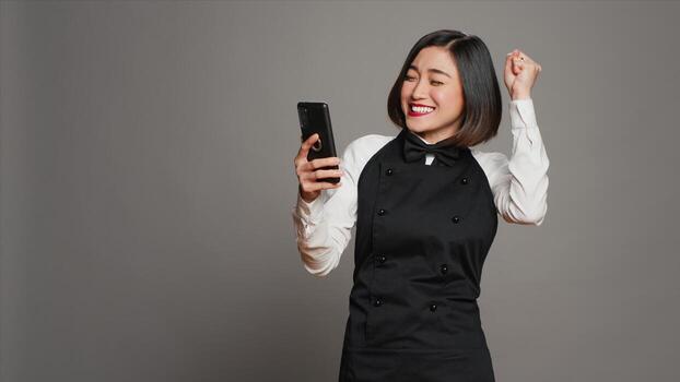 Asian server feeling happy after reading good news on phone, having a lot of clients with reservations for dinner. Waitress with apron using smartphone to see table bookings, enjoys work. Camera A. photo