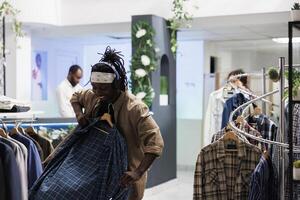 Happy shopper listening to music while choosing trendy shirt to buy in clothing store. Cheerful smiling african american man in headphones holding apparel on hanger in shopping mall photo