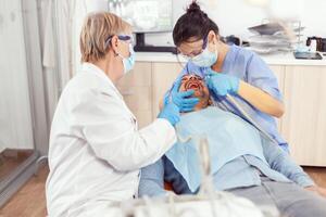 enfermo hombre sentado en médico silla con abierto boca mientras médico enfermero con cara máscara y guantes limpieza dientes después dental cirugía. hospital equipo trabajando en estomatológico clínica foto