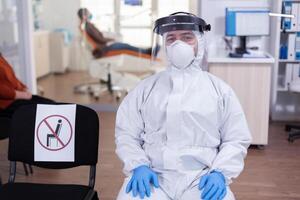 Close up of tired doctor in stomatological office looking on camera wearing overall and face shield sitting on chair in waiting room clinic. Concept of new normal dentist visit in coronavirus outbreak. photo