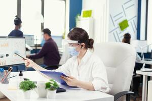 Manager wearing protection against covid 19 during global pandemic. Employees with visors working in corporate company workspace respecting social distance analysing data and graphs. photo