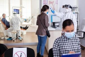 Stomatology receptionist using digital thermometer to measure patient temperature dressed in ppe suit as safety precaution during global outreabk with coronavirus. Dentist treating teeth. photo