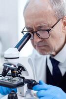 Elderly aged scientist in modern facility looking through a microscope during health care study. Chemist researcher in sterile lab doing experiments for medical industry using modern technology. photo