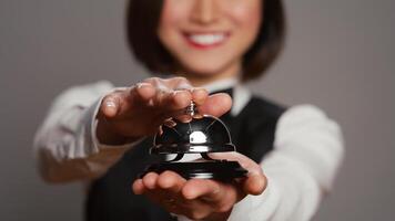 Receptionist posing with service bell and ringing it on camera, expressing assistance from front desk staff. Asian hotel concierge with hospitality industry operator occupation. Close up. Camera B. photo