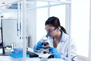 Scientist in laboratory doing research looking through microscope wearing protection glasses and gloves. Chemist wearing lab coat using modern technology during scientific experiment in sterile environment. photo