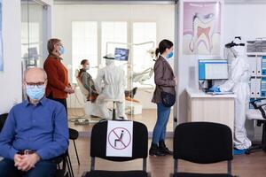 Patients with protection mask waiting in stomatology reception with new normal, staff wearing ppe suit. Woman checking appoiment with nurse coming for dental control during coronavirus pandemic photo