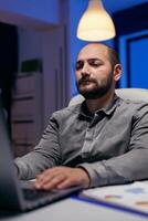 Serious young manager working on deadline doing overtime in empty office. Smart businessman sitting at his workplace in the course of late night hours doing his job. photo