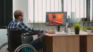 Handicapped game developer sitting in wheelchair with protection mask working at new project from new normal studio office during covid-19 pandemic. Immobilized man respecting social distance. photo