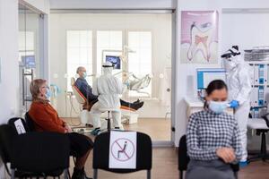 Doctor in pee suit consulting senior patient in stomatology office keeping visor, during global pandemic with coronavirus, new normal. Receptionist with face mask in dentistiry waiting area. photo