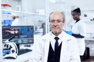 mayor científico sentado a su lugar de trabajo durante médico experimentos mayor científico vistiendo laboratorio Saco trabajando a desarrollar un nuevo médico vacunar con africano asistente en el antecedentes. foto