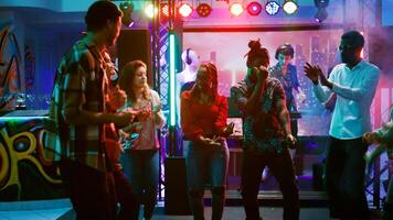 Young adults dancing at nightclub and enjoying electronic music at dance party. Modern people dancing and partying together on funky beats, having fun with stage lights. Handheld shot. photo