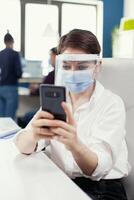 Entrepreneur wearing face mask as safety precaution against covid 19 chatting on smartphone at workplace.Multiethnic coworkers working respecting social distance in financial company. New normal. photo