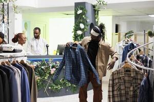 Shopper enjoying music in headphones while browsing for trendy clothes in mall. Showroom customer in earphones holding casual plaid shirt on hanger to try on in fitting room photo