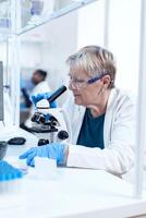 Senior scientist woman with protection glasses doing genetic investigation using microscope with african assistant in the background.. Elderly researcher carrying out scientific research in a sterile lab using a modern technology. photo