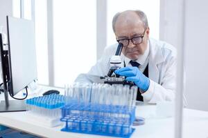 Healthcare senior scientist man in medical industry carrying out genetic analysis using microscope. Chemist researcher in sterile lab doing experiments for medical industry using modern technology. photo