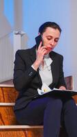 Overworked tired businesswoman reading project deadline during phone call with coworker sitting on staircase of business building. Serious entrepreneur working on corporate job late at night. photo