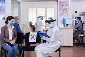 Dentist assistant with ppe equipment talking with patient before consultation during coronavirus epidemic sitting on chairs in waiting area keeping distance. Concept of new normal dentist visit. photo