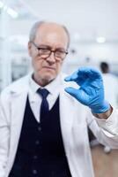 Concentrated senior scientist looking at glass slide with sample wearing protection gloves. Elderly researcher in sterile lab looking on microscope slide wearing lab coat. photo