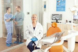 Portrait of senior dentist woman sitting on chair in stomatological hospital office. In background dentist assistant talking with sick patient for dental treatment during stomatology consultation photo