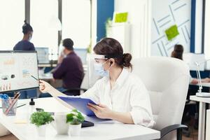 enfocado mujer de negocios en un remoto llamada utilizando inalámbrico auriculares vistiendo cara máscara en contra COVID-19. empleados con viseras trabajando en corporativo empresa espacio de trabajo respecto a social distancia analizando datos y gráficos foto