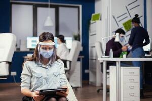 Woman entrepreneur with protection mask against covid-19 pandemic sitting in middle of office room with new normal holding digital tablet. Multiethnic business team working respecting social distance during global pandemic with covid-19. photo