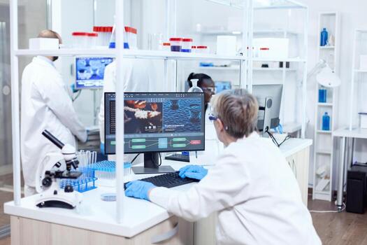 Elderly researcher engineering human adn using softtware on powerfull computer. Senior scientist in pharmaceuticals laboratory doing genetic research wearing lab coat with team in the background. photo