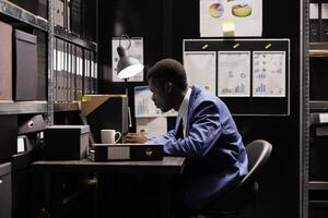 Depository employee working at bureaucracy research, analyzing administrative documents in arhive room. African american corporate worker discovering files with accountancy record photo